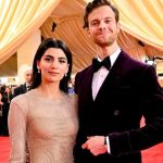 Jack Quaid and Claudia Doumit pose together on a red carpet at a formal event. Claudia wears a shimmering, long-sleeved neutral-toned gown, while Jack wears a deep purple velvet tuxedo with a black bow tie against a backdrop of gold draped curtains.