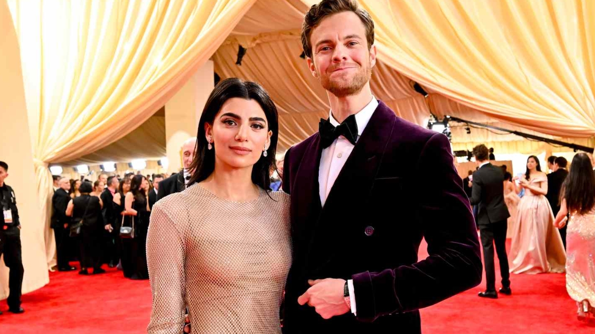 Jack Quaid and Claudia Doumit pose together on a red carpet at a formal event. Claudia wears a shimmering, long-sleeved neutral-toned gown, while Jack wears a deep purple velvet tuxedo with a black bow tie against a backdrop of gold draped curtains.