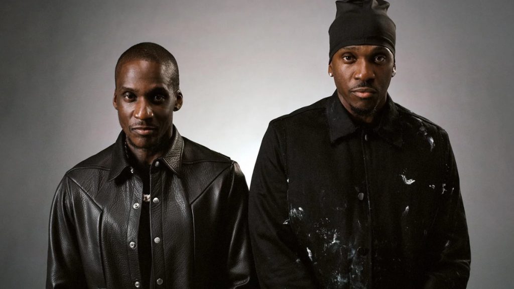 A professional studio portrait of the hip-hop duo Clipse, featuring brothers Pusha T and No Malice. Both are dressed in black, with No Malice wearing a leather jacket and Pusha T in a black denim jacket and a headwrap.