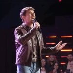 Dylan Carter , performing on a brightly lit stage. He is wearing a brown leather jacket over a button-down shirt and light-wash jeans, singing into a handheld microphone with an emotional expression. In the background, a studio audience is visible beneath horizontal red stage lights.