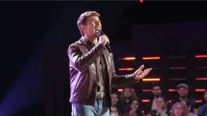 Dylan Carter , performing on a brightly lit stage. He is wearing a brown leather jacket over a button-down shirt and light-wash jeans, singing into a handheld microphone with an emotional expression. In the background, a studio audience is visible beneath horizontal red stage lights.
