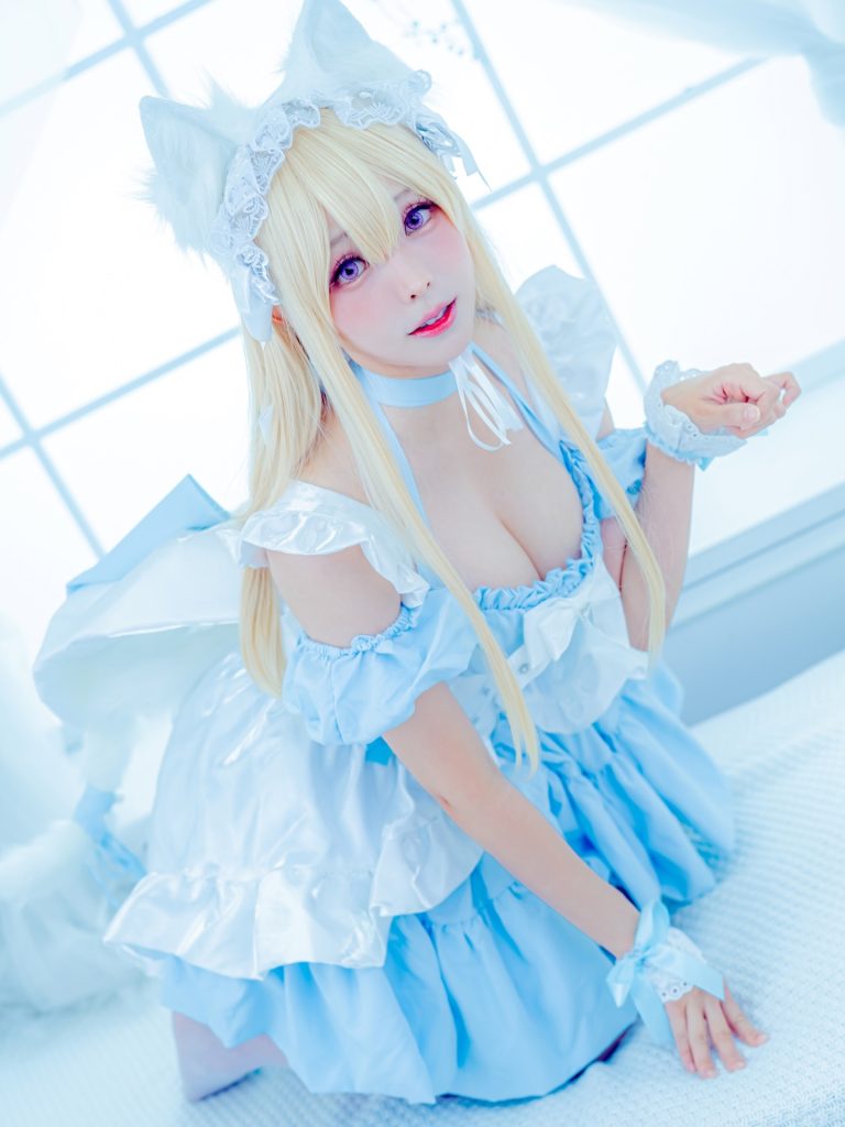 Ely Cosplay cosplaying as a character in an intricate blue and white outfit with floral patterns and gold accents. She is holding a traditional-style fan, and the background features soft, out-of-focus indoor lighting.