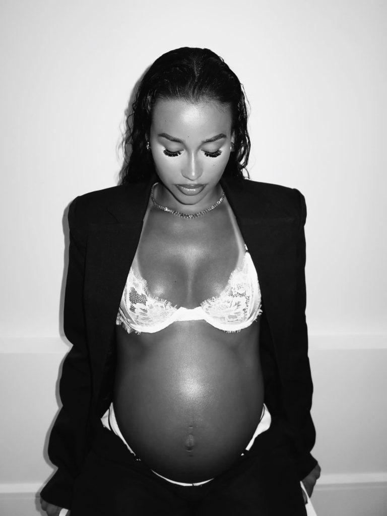 A black and white maternity photograph of Jada Wallace. She is looking down at her pregnant belly, wearing an open black blazer over a white lace bra.