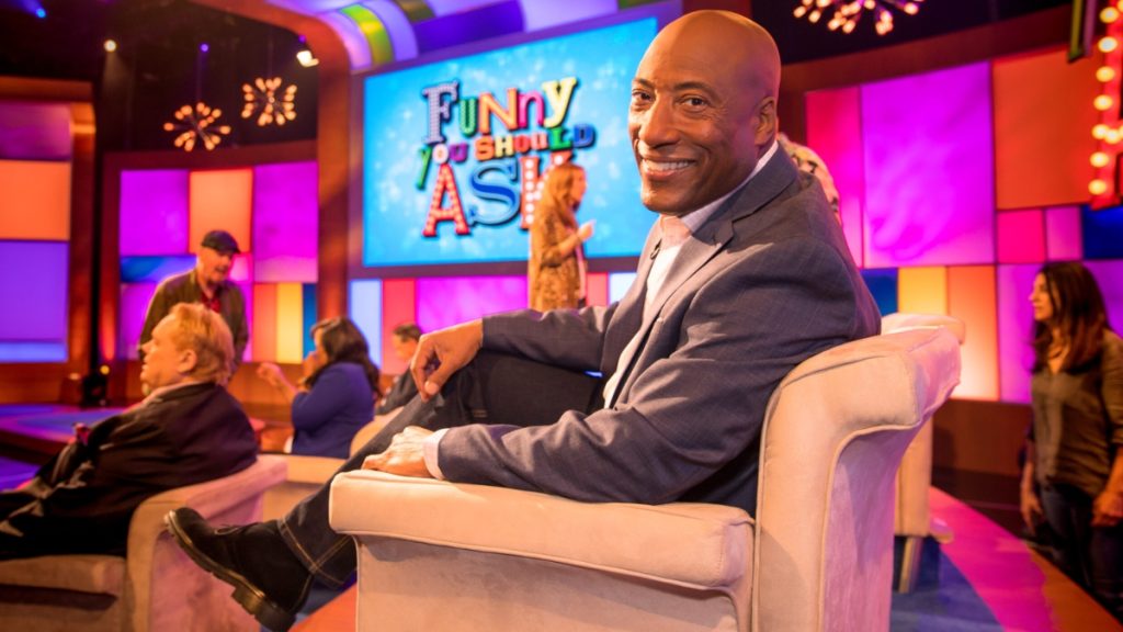 Byron Allen sits in a light-colored armchair on the vibrant, colorful set of the game show "Funny You Should Ask." He is wearing a blue suit jacket and smiling at the camera, with the show's logo visible on a large screen in the background.