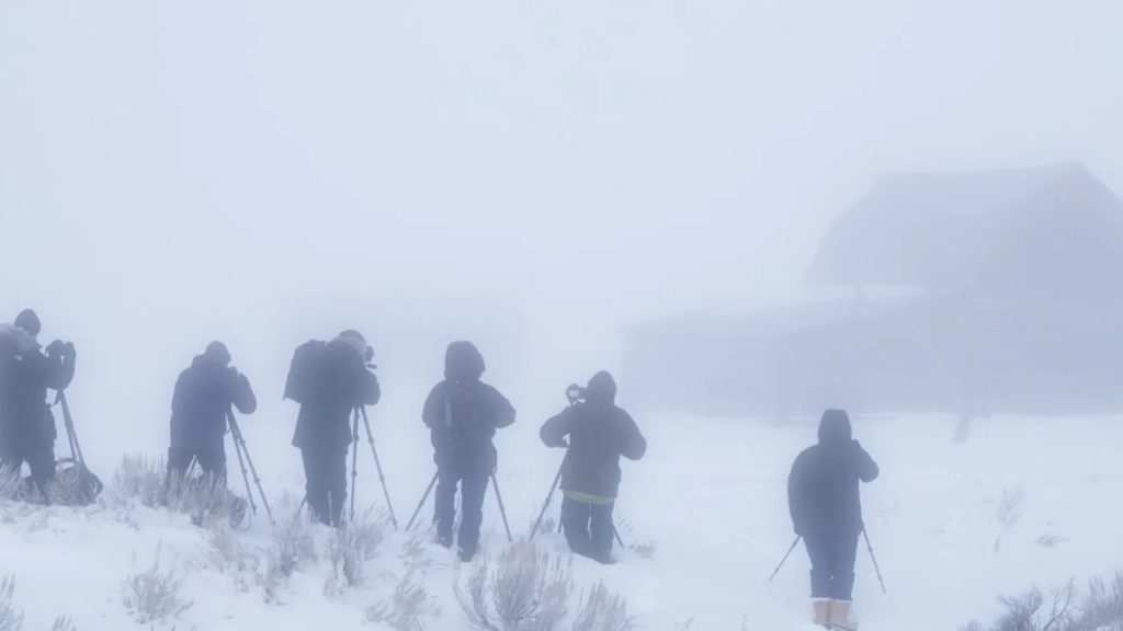 A group of photographers with tripods and heavy winter gear stand in a whiteout during a Heavy Snow Warning. The scene is obscured by thick fog and falling snow, with a faint silhouette of a building in the background, illustrating the dangerous visibility and extreme conditions of a major winter storm.
