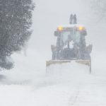 A large yellow snow plow clears a road during a Heavy Snow Warning. Thick snow is falling, and the plow’s lights are visible through the haze as it pushes through deep accumulations. The surrounding trees are heavily coated in white, highlighting the intensity of the ongoing blizzard.