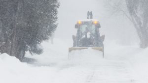 A large yellow snow plow clears a road during a Heavy Snow Warning. Thick snow is falling, and the plow’s lights are visible through the haze as it pushes through deep accumulations. The surrounding trees are heavily coated in white, highlighting the intensity of the ongoing blizzard.