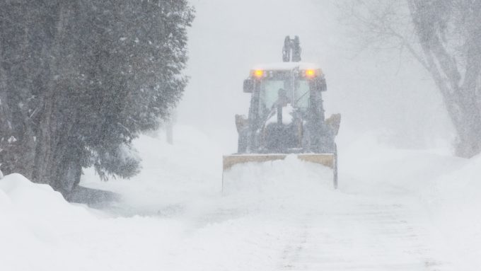 A large yellow snow plow clears a road during a Heavy Snow Warning. Thick snow is falling, and the plow’s lights are visible through the haze as it pushes through deep accumulations. The surrounding trees are heavily coated in white, highlighting the intensity of the ongoing blizzard.