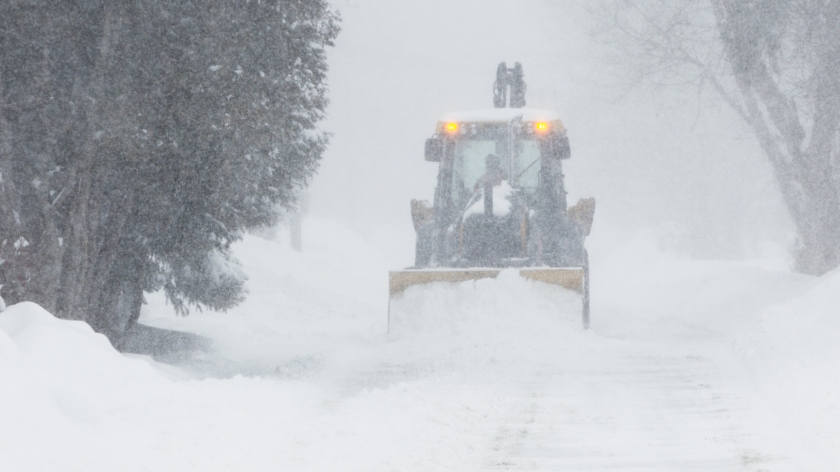 A large yellow snow plow clears a road during a Heavy Snow Warning. Thick snow is falling, and the plow’s lights are visible through the haze as it pushes through deep accumulations. The surrounding trees are heavily coated in white, highlighting the intensity of the ongoing blizzard.
