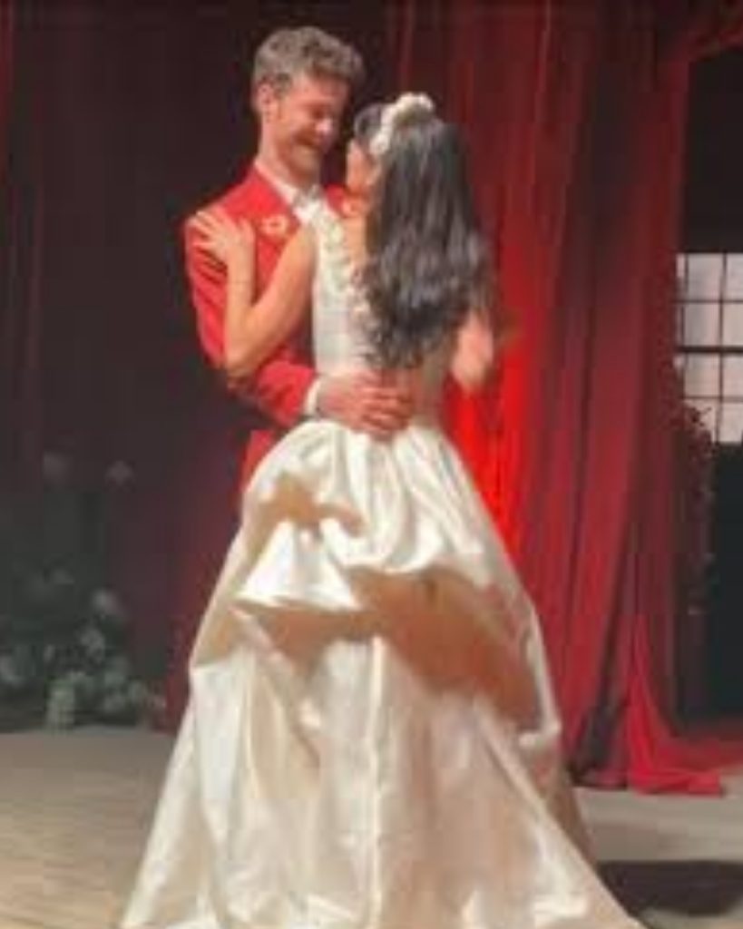 An intimate photo from the wedding of Jack Quaid and Claudia Doumit in Australia. Claudia is seen from the back in a classic white satin wedding gown with a voluminous skirt and floral headband, while Jack smiles at her wearing a vibrant red suit jacket.