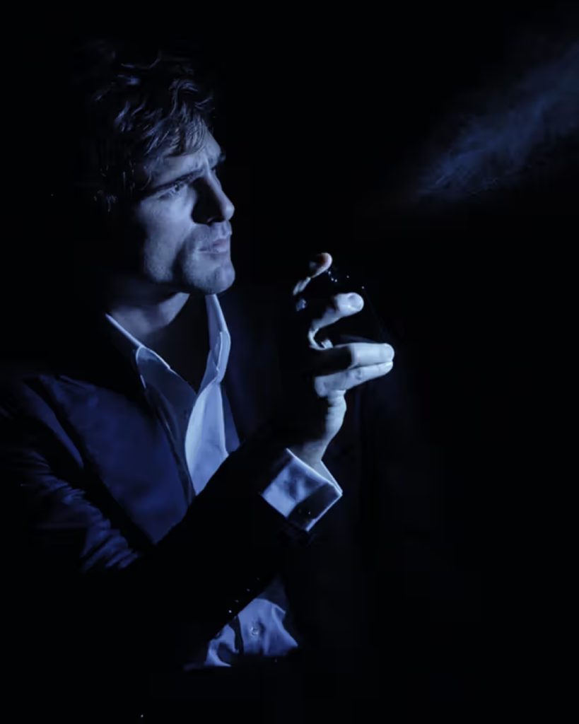 A moody, low-light photograph of Jacob Elordi Chanel ambassador holding a fragrance bottle. He is dressed in a dark suit and a crisp white shirt, captured in profile as he sprays the cologne. A fine mist of the scent is visible, illuminated against the dark background by a cool blue light source.