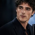 A promotional campaign image for Jacob Elordi Chanel featuring the actor looking off-camera with a contemplative expression. He is wearing a sharp black suit and a matching black button-down shirt. In the foreground, a large bottle of "Bleu de Chanel L'Exclusif" is prominently displayed, with the interlocking Chanel "CC" logo blurred in the deep blue background.