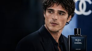 A promotional campaign image for Jacob Elordi Chanel featuring the actor looking off-camera with a contemplative expression. He is wearing a sharp black suit and a matching black button-down shirt. In the foreground, a large bottle of "Bleu de Chanel L'Exclusif" is prominently displayed, with the interlocking Chanel "CC" logo blurred in the deep blue background.