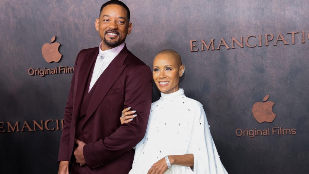 Will Smith and Jada Pinkett Smith posing together on a red carpet for the film "Emancipation." Will is wearing a maroon three-piece suit, and Jada is in a high-neck white gown with a flowing cape. The "Apple Original Films" logo is visible on the backdrop behind them.