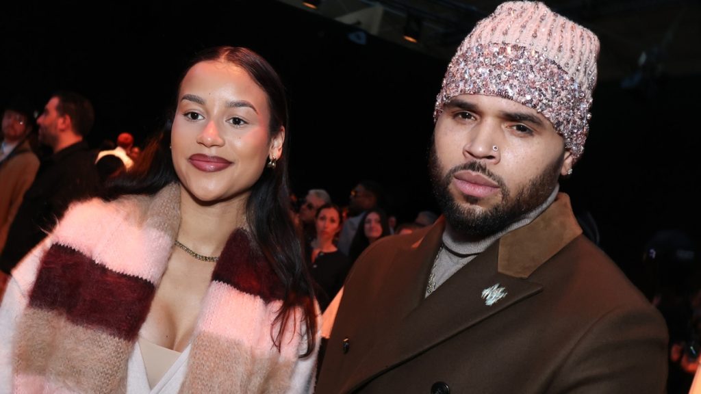 A close-up shot of Jada Wallace and Chris Brown posing together at a crowded event. Jada is wearing a multi-colored pink and tan fuzzy scarf and smiling slightly. Chris Brown wears a brown coat over a turtleneck and a pink, sequin-covered beanie.