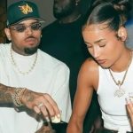 Chris Brown and Jada Wallace sitting together at a table during a social gathering. Chris Brown is wearing a white t-shirt, a green "VIB" baseball cap, and jeweled sunglasses while leaning over to pour a drink. Jada Wallace wears a white tank top and a gold "OHB" pendant necklace, looking down as she holds a shot glass.