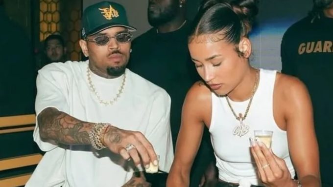Chris Brown and Jada Wallace sitting together at a table during a social gathering. Chris Brown is wearing a white t-shirt, a green "VIB" baseball cap, and jeweled sunglasses while leaning over to pour a drink. Jada Wallace wears a white tank top and a gold "OHB" pendant necklace, looking down as she holds a shot glass.