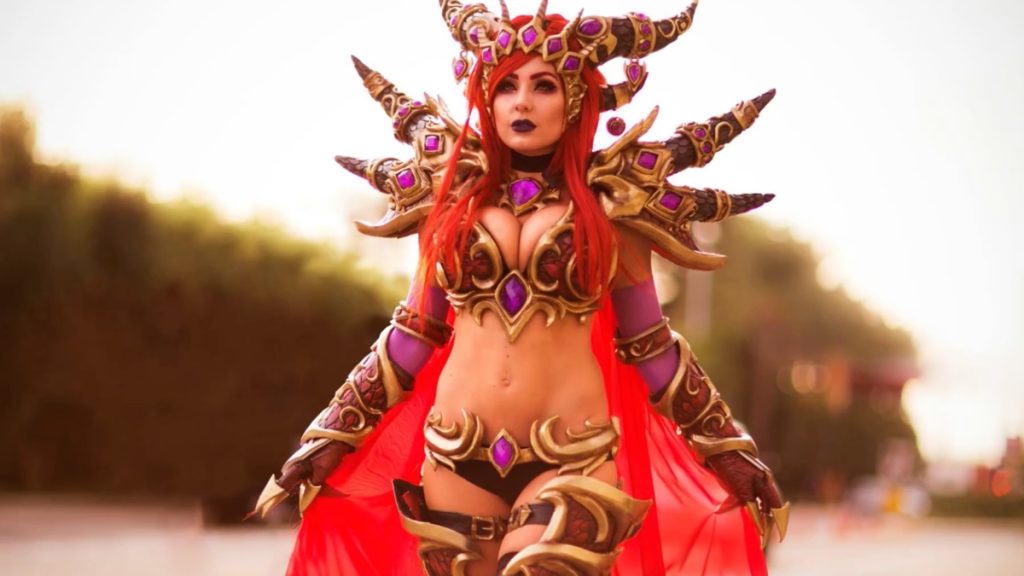 An outdoor, cinematic shot of Jessica Nigri in an elaborate Alexstrasza the Life-Binder cosplay. The costume features intricate, dark-gold dragon-scaled armor with purple glowing gems, a dramatic horned headpiece, and a flowing red cape. Her red hair and dark makeup contrast against a warm, blurred sunset background.