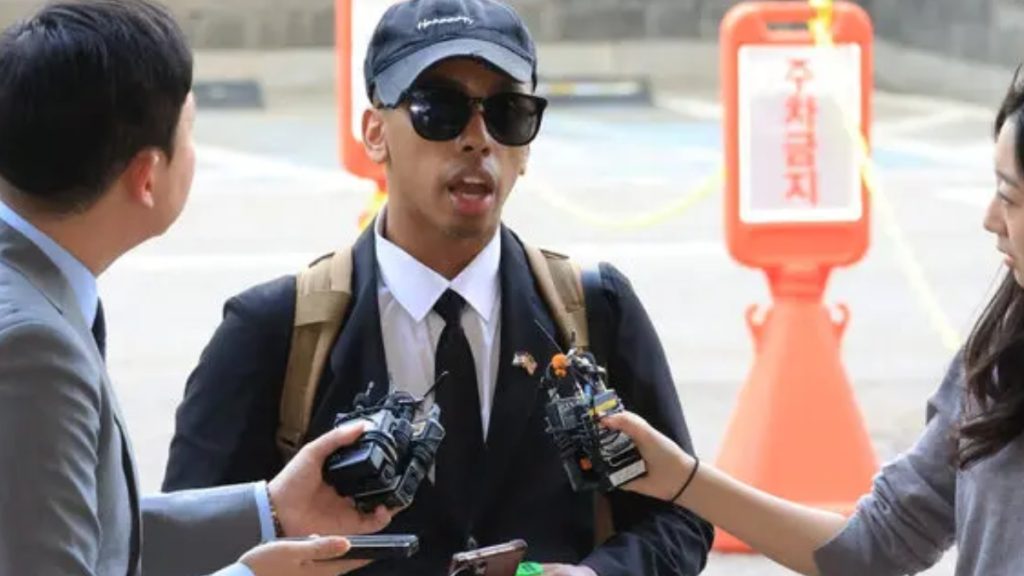 A medium shot of Johnny Somali being interviewed by reporters. He is dressed formally in a black suit, white dress shirt, and black tie, though he is also wearing a black baseball cap and dark sunglasses. He has a tan backpack on his shoulders. Several hands holding microphones and recording devices are extended toward him from the left and right. In the background, a bright orange traffic pylon with Korean text is visible on the pavement.