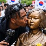 A medium shot captures live-streamer Johnny Somali leaning in to kiss the cheek of the "Statue of Peace" in South Korea. He is a young Black man with short dreadlocks and sunglasses perched on his head, wearing a black hoodie and holding a camera rig. The bronze statue depicts a seated young woman in traditional Korean hanbok, symbolizing "comfort women" victims. In the blurred background, a crowd of people and several South Korean flags are visible, with some individuals appearing to be shouting or protesting.