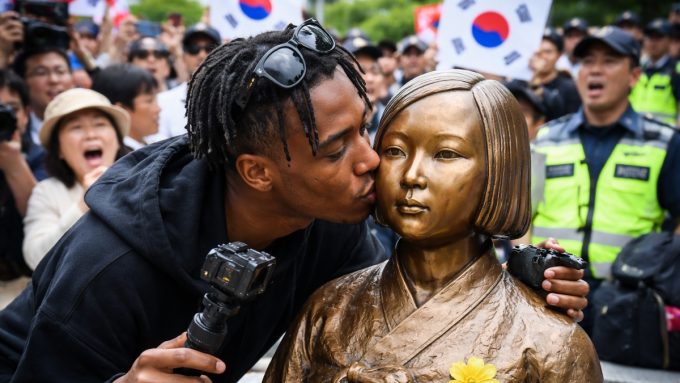 A medium shot captures live-streamer Johnny Somali leaning in to kiss the cheek of the "Statue of Peace" in South Korea. He is a young Black man with short dreadlocks and sunglasses perched on his head, wearing a black hoodie and holding a camera rig. The bronze statue depicts a seated young woman in traditional Korean hanbok, symbolizing "comfort women" victims. In the blurred background, a crowd of people and several South Korean flags are visible, with some individuals appearing to be shouting or protesting.