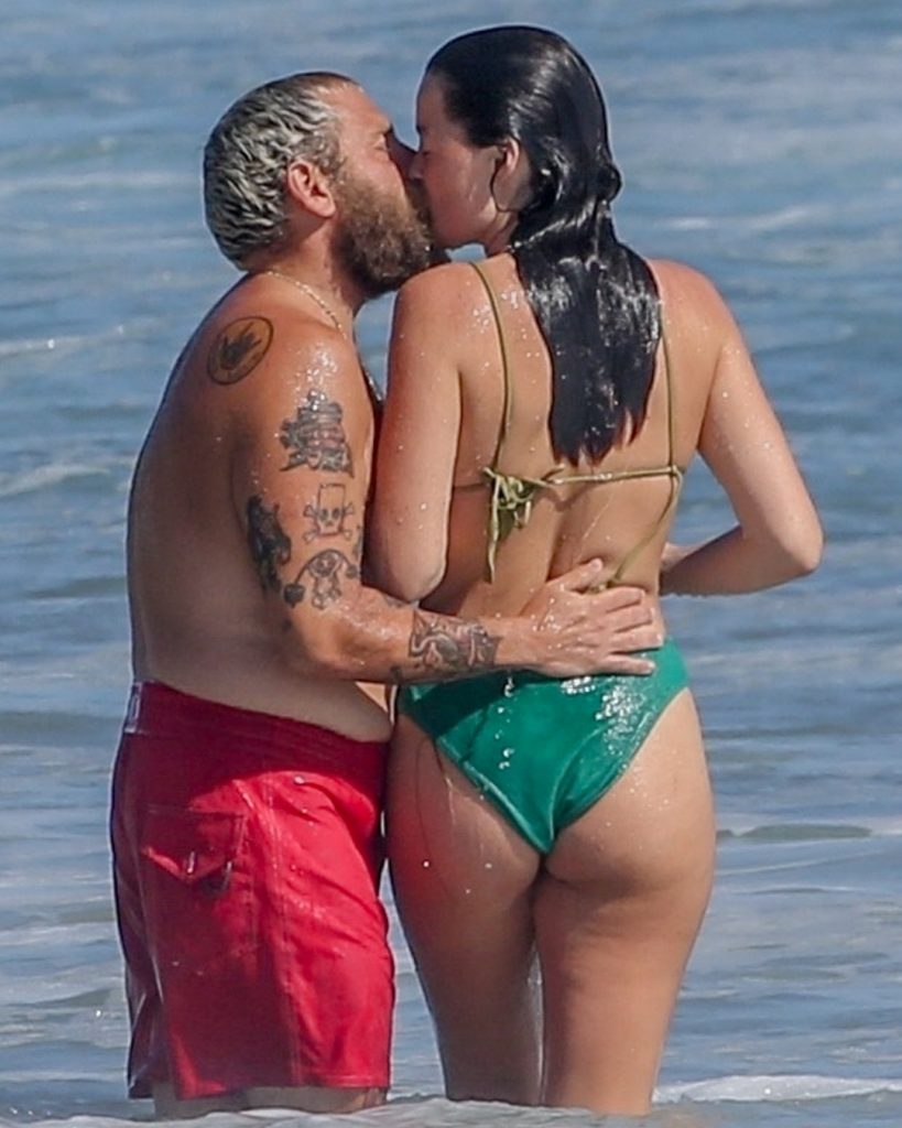A candid photo of Jonah Hill and Olivia Millar sharing a kiss while standing in the ocean. Jonah is wearing red swim trunks, and Olivia is in a green bikini.