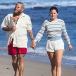 Jonah Hill and Olivia Millar walking hand-in-hand along a sandy beach with the ocean waves in the background. Jonah is wearing an open white button-down shirt and red shorts, while Olivia wears a striped long-sleeve top and white shorts.
