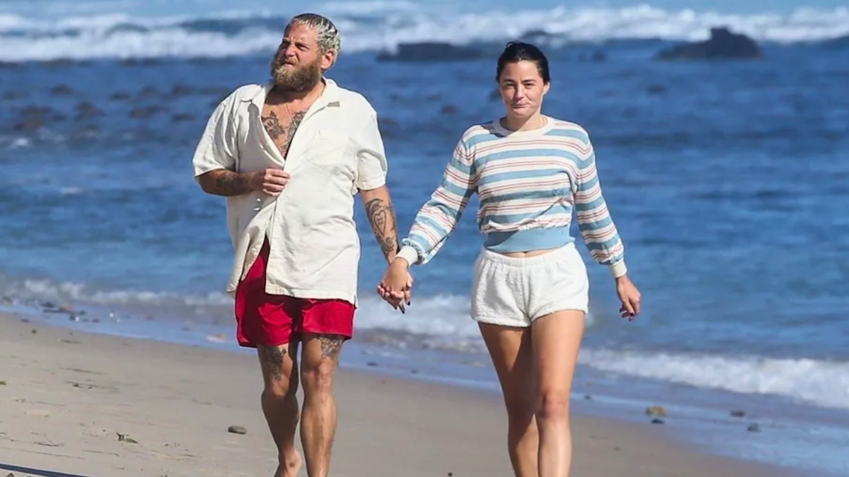 Jonah Hill and Olivia Millar walking hand-in-hand along a sandy beach with the ocean waves in the background. Jonah is wearing an open white button-down shirt and red shorts, while Olivia wears a striped long-sleeve top and white shorts.