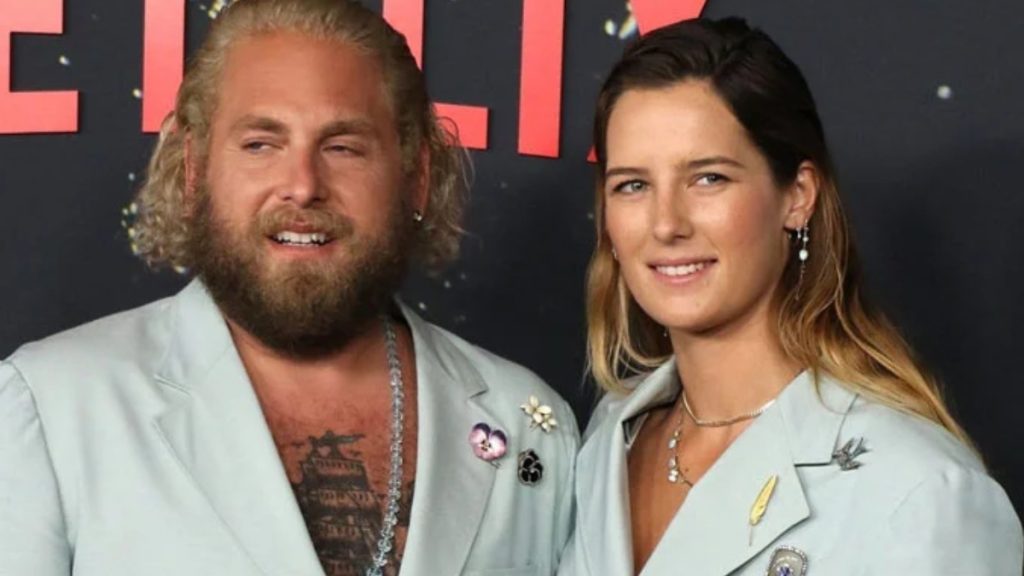 Actor Jonah Hill and Olivia Millar posing together at a red carpet event. They are wearing matching light blue oversized blazers. Jonah has a full beard and blonde hair, while Olivia has long brown hair and is wearing delicate layered necklaces.