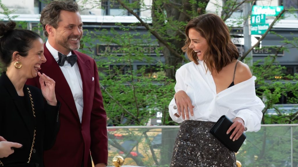 A candid medium shot of Joshua Jackson and Katie Holmes sharing a laugh at an outdoor event in New York City. Joshua is dressed in a burgundy tuxedo jacket with a black bow tie, looking over at Katie with a wide smile. Katie is laughing heartily, wearing an off-the-shoulder white button-down shirt paired with a shimmering silver sequined skirt. A third woman in a black blazer is partially visible on the left, also laughing. Green trees and city street signs are visible in the background.