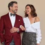 A medium shot of Joshua Jackson and Katie Holmes posing together against a neutral, tan-colored backdrop. Katie is linked arm-in-arm with Joshua, leaning toward him and laughing. Joshua, in his burgundy tuxedo, looks at her with a warm, smiling expression. Katie’s outfit—the white shirt falling off one shoulder and the textured grey-silver skirt—is shown in full detail as they recreate their famous chemistry for the cameras.