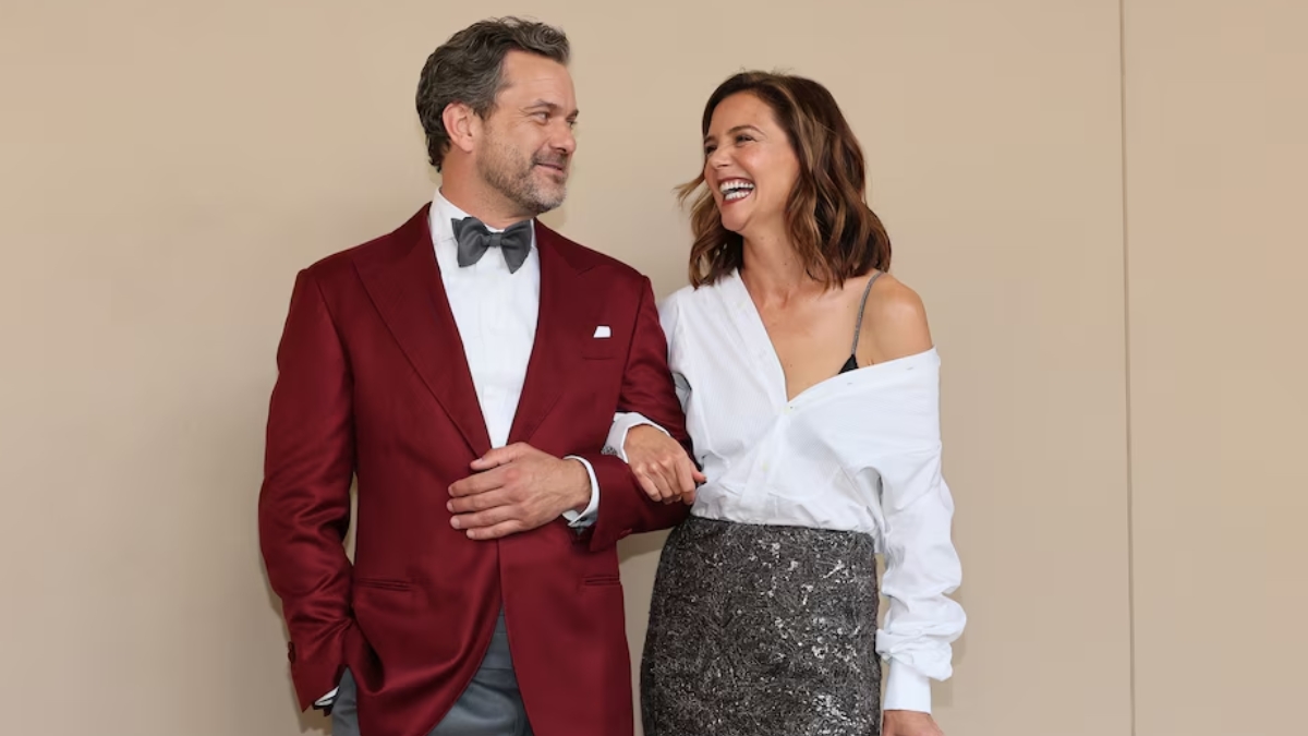 A medium shot of Joshua Jackson and Katie Holmes posing together against a neutral, tan-colored backdrop. Katie is linked arm-in-arm with Joshua, leaning toward him and laughing. Joshua, in his burgundy tuxedo, looks at her with a warm, smiling expression. Katie’s outfit—the white shirt falling off one shoulder and the textured grey-silver skirt—is shown in full detail as they recreate their famous chemistry for the cameras.