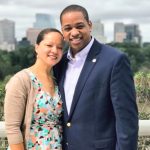 A smiling Justin Fairfax and his wife, Dr. Cerina Fairfax, stand closely together outdoors. Justin, a former Virginia Lieutenant Governor, is wearing a blue blazer over a light-colored button-down shirt. Cerina is wearing a colorful geometric-patterned wrap dress with a tan cardigan. They are posed in front of a white railing with a city skyline and lush green trees visible in the blurred background under an overcast sky.