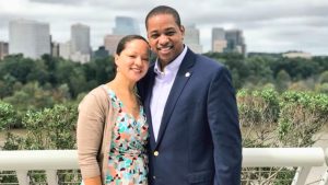 A smiling Justin Fairfax and his wife, Dr. Cerina Fairfax, stand closely together outdoors. Justin, a former Virginia Lieutenant Governor, is wearing a blue blazer over a light-colored button-down shirt. Cerina is wearing a colorful geometric-patterned wrap dress with a tan cardigan. They are posed in front of a white railing with a city skyline and lush green trees visible in the blurred background under an overcast sky.
