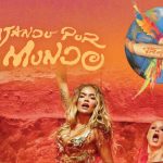 Promotional image for the Karol G concert tour titled "Viajando Por El Mundo Tropitour." The graphic features Karol G performing on stage in a gold fringed outfit alongside backup dancers against a warm orange backdrop. Stylized text reads "Viajando Por El Mundo" next to a logo of a colorful macaw perched on a globe wrapped in a banner that says "Tropitour."
