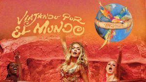 Promotional image for the Karol G concert tour titled "Viajando Por El Mundo Tropitour." The graphic features Karol G performing on stage in a gold fringed outfit alongside backup dancers against a warm orange backdrop. Stylized text reads "Viajando Por El Mundo" next to a logo of a colorful macaw perched on a globe wrapped in a banner that says "Tropitour."