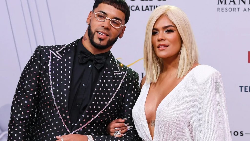 A red carpet photo of Karol G and Anuel AA from their relationship timeline. Anuel AA is wearing a black suit with silver stud detailing and white-rimmed glasses. Karol G is posing beside him with platinum blonde hair, wearing a white, deep-V sequined gown.