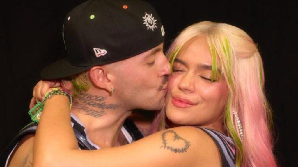 A candid, close-up photo of Karol G and Feid. Feid, wearing a black baseball cap, is leaning in to kiss Karol G on the cheek while she smiles with her eyes closed. Karol G has pink hair with neon green highlights and a visible heart tattoo on her shoulder.