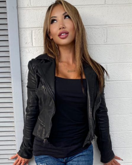 Kiara Moon in black leather jacket, t-shirt, and ripped blue jeans, posing.