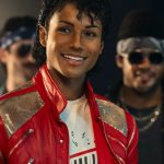 A production still from the Michael Movie featuring Jaafar Jackson as Michael Jackson. He is smiling brightly, wearing an iconic red leather jacket with silver metallic panels over a graphic t-shirt. He has Michael’s signature jheri curl hairstyle and is surrounded by a group of background dancers in a dimly lit studio setting.