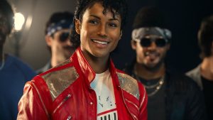 A production still from the Michael Movie featuring Jaafar Jackson as Michael Jackson. He is smiling brightly, wearing an iconic red leather jacket with silver metallic panels over a graphic t-shirt. He has Michael’s signature jheri curl hairstyle and is surrounded by a group of background dancers in a dimly lit studio setting.