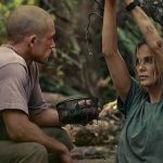 A dramatic still from the Apex Movie featuring Taron Egerton and Charlize Theron in a wooded, outdoor setting. Taron Egerton, with a shaved head and wearing a tan t-shirt, is crouched on the left, holding a small wire basket. Charlize Theron is on the right, leaning against a tree with her arms raised and bound by rope to a branch. Both characters appear weathered and dirty, looking at each other with intense expressions.