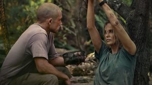 A dramatic still from the Apex Movie featuring Taron Egerton and Charlize Theron in a wooded, outdoor setting. Taron Egerton, with a shaved head and wearing a tan t-shirt, is crouched on the left, holding a small wire basket. Charlize Theron is on the right, leaning against a tree with her arms raised and bound by rope to a branch. Both characters appear weathered and dirty, looking at each other with intense expressions.