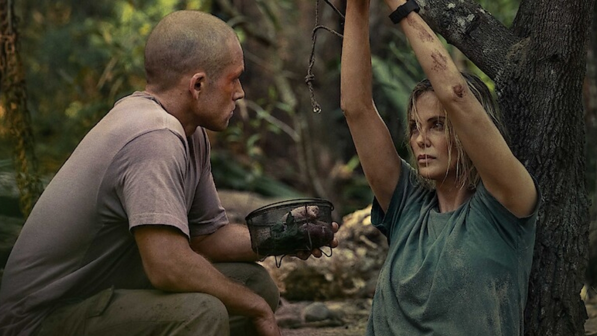 A dramatic still from the Apex Movie featuring Taron Egerton and Charlize Theron in a wooded, outdoor setting. Taron Egerton, with a shaved head and wearing a tan t-shirt, is crouched on the left, holding a small wire basket. Charlize Theron is on the right, leaning against a tree with her arms raised and bound by rope to a branch. Both characters appear weathered and dirty, looking at each other with intense expressions.