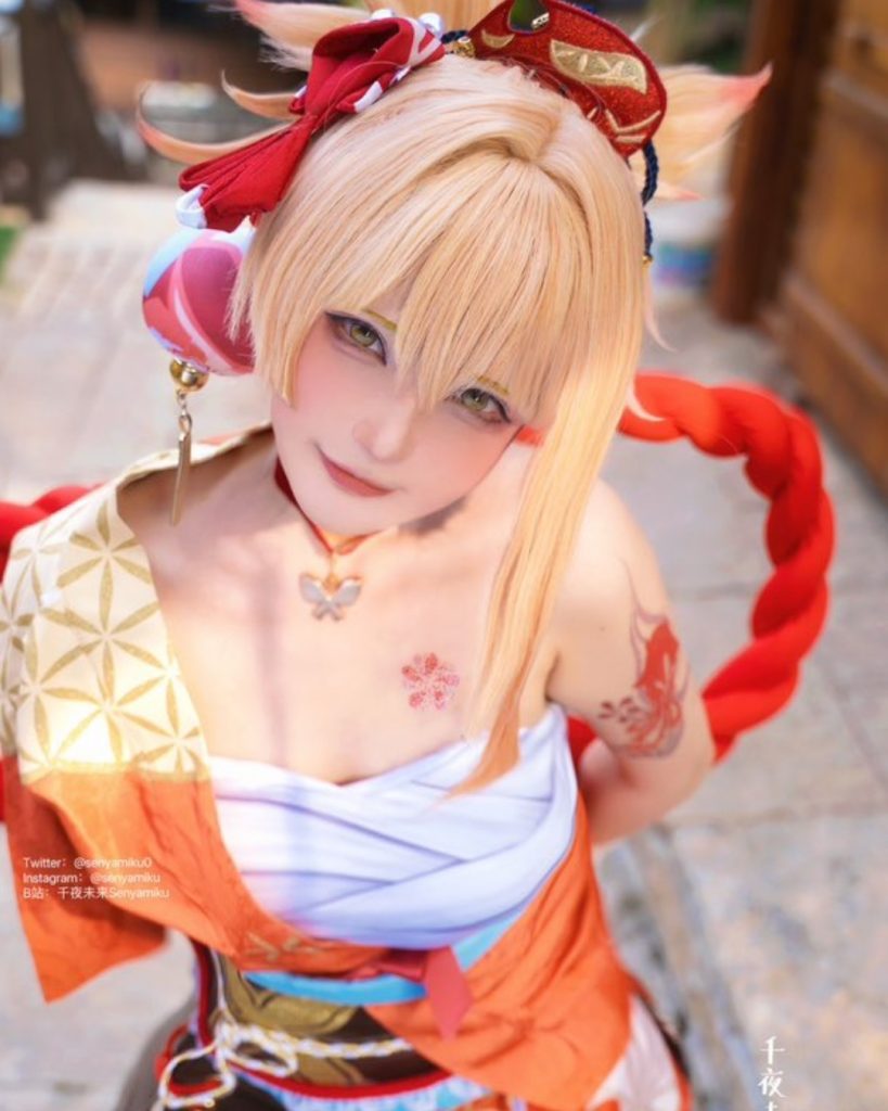 Senyamiku cosplaying as Yoimiya from Genshin Impact. She is seen from a high angle, featuring her blonde hair styled with red ribbons and a floral tattoo on her shoulder. She is wearing a traditional-inspired orange and white outfit with a large red decorative rope behind her.