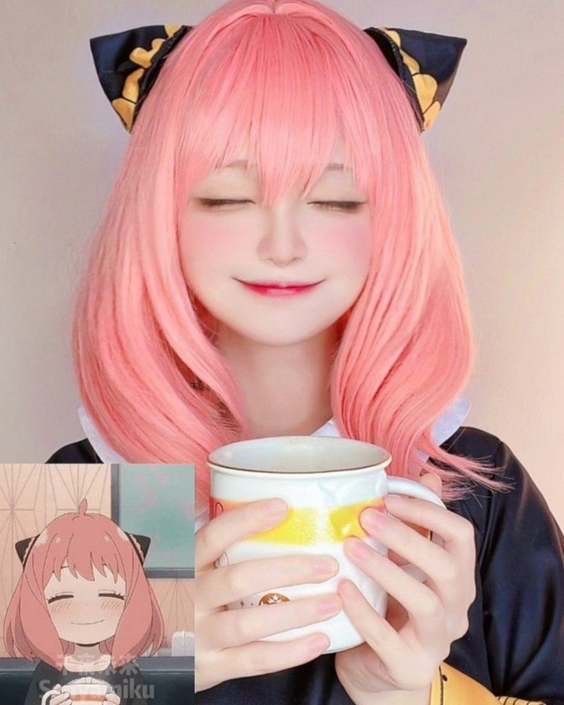 Senyamiku cosplaying as Anya Forger from Spy × Family. She has pastel pink hair with black and gold hair ornaments, her eyes are closed in a smile as she holds a large white and yellow mug. A small reference image of the anime character is in the bottom left corner.