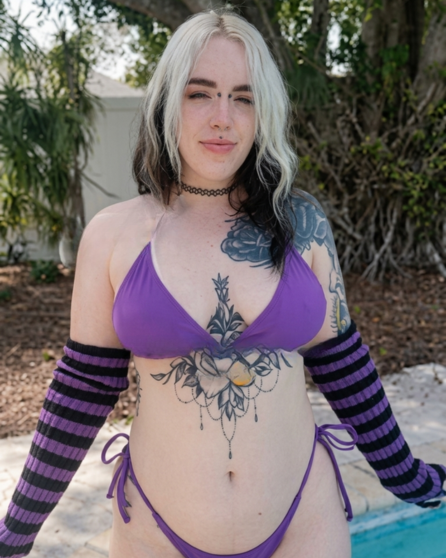 Valerie Vampyr posing in a purple string bikini with tattoos by the pool.