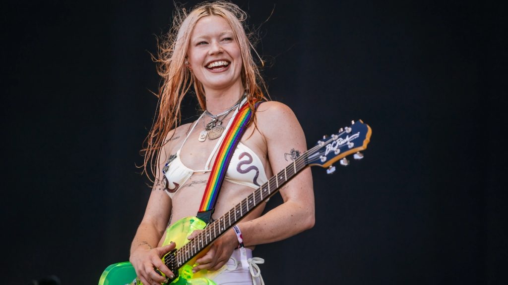 Rhian Teasdale of the indie rock band Wet Leg performing live on stage. She is smiling while playing a neon green electric guitar with a rainbow strap, wearing a white bikini top and silver necklaces.