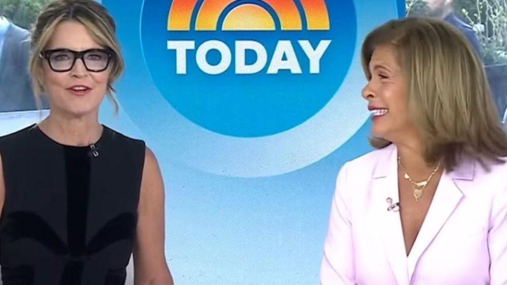 A bright studio shot from the Today show featuring Savannah Guthrie and Hoda Kotb in front of the iconic colorful sun logo. On the left, Savannah Guthrie wears black-rimmed glasses and a black sleeveless top, looking toward the camera. On the right, Hoda Kotb is shown in profile wearing a light pink blazer and gold necklaces, smiling warmly. This image is often associated with discussions regarding when savannah guthrie leaves today show early or handles unexpected broadcast changes.