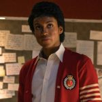 A production still featuring Jaafar Jackson portraying Michael Jackson. He is standing in a room with a wall covered in handwritten notes and papers. He wears a red cardigan with a "J" crest and white stripes on the sleeve over a white collared shirt, sporting a signature curly hairstyle.